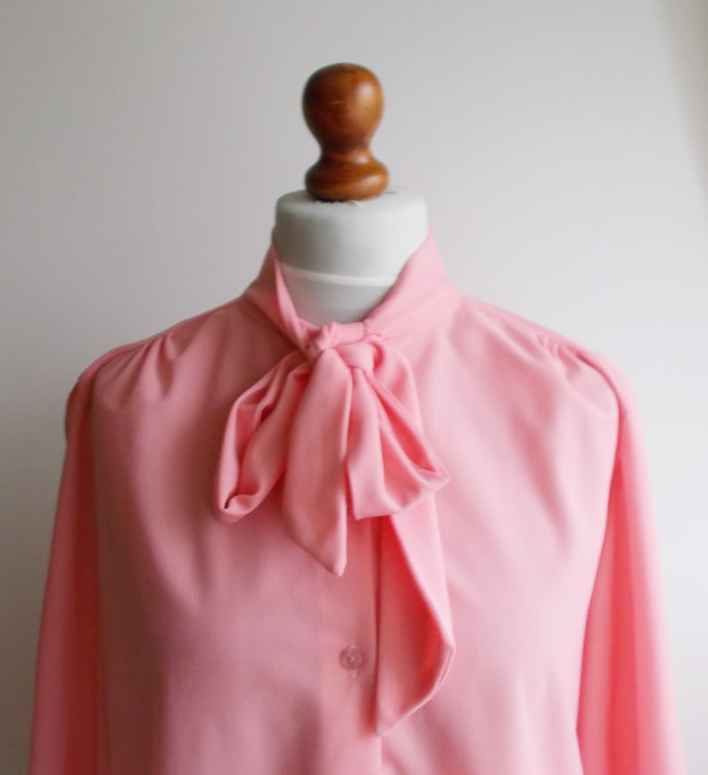 May include: A pink blouse with a large bow tie at the neck. The blouse has long sleeves and a button-up front.