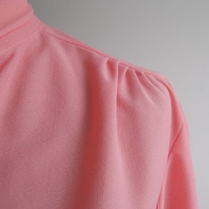May include: Close-up of a pink, textured blouse with a gathered detail at the shoulder.