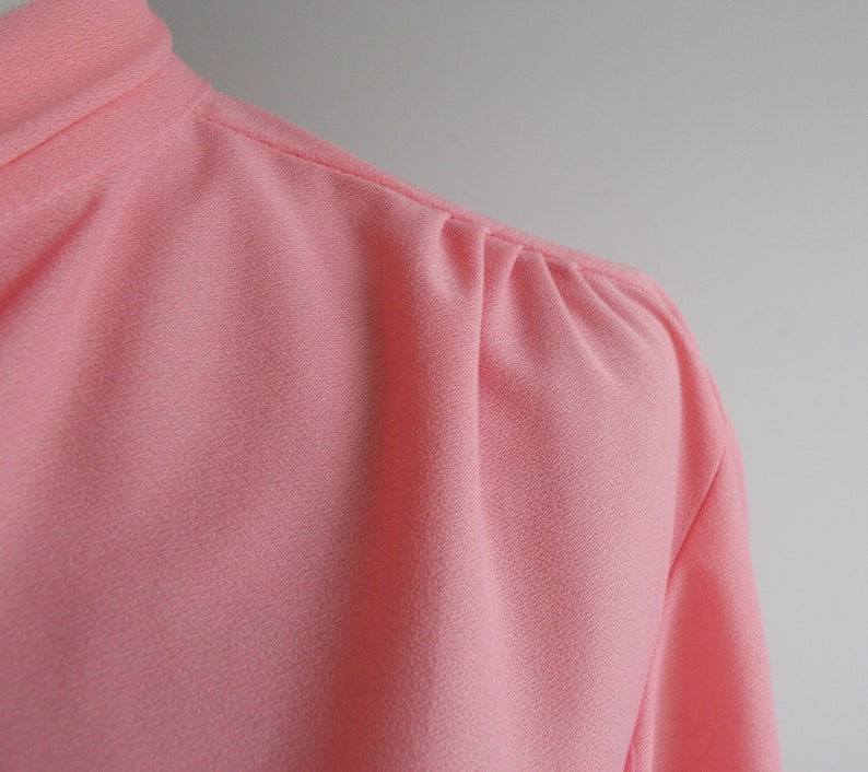 May include: Close-up of a pink, textured blouse with a gathered detail at the shoulder.