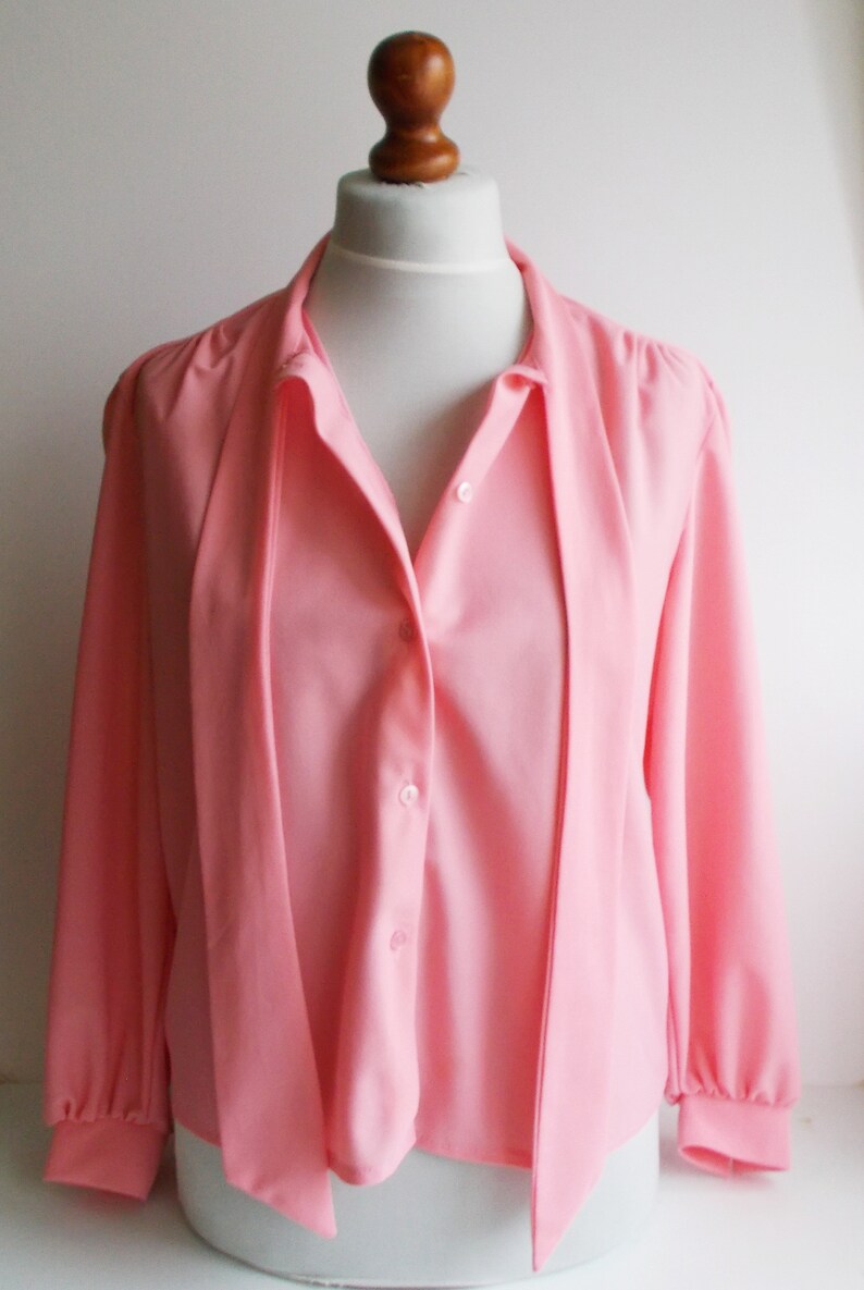 May include: A pink long-sleeved blouse with a tie at the neck and button closure.