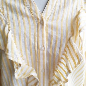 May include: A white and yellow striped button-up shirt with ruffled detailing. The shirt has a v-neck and a button closure.