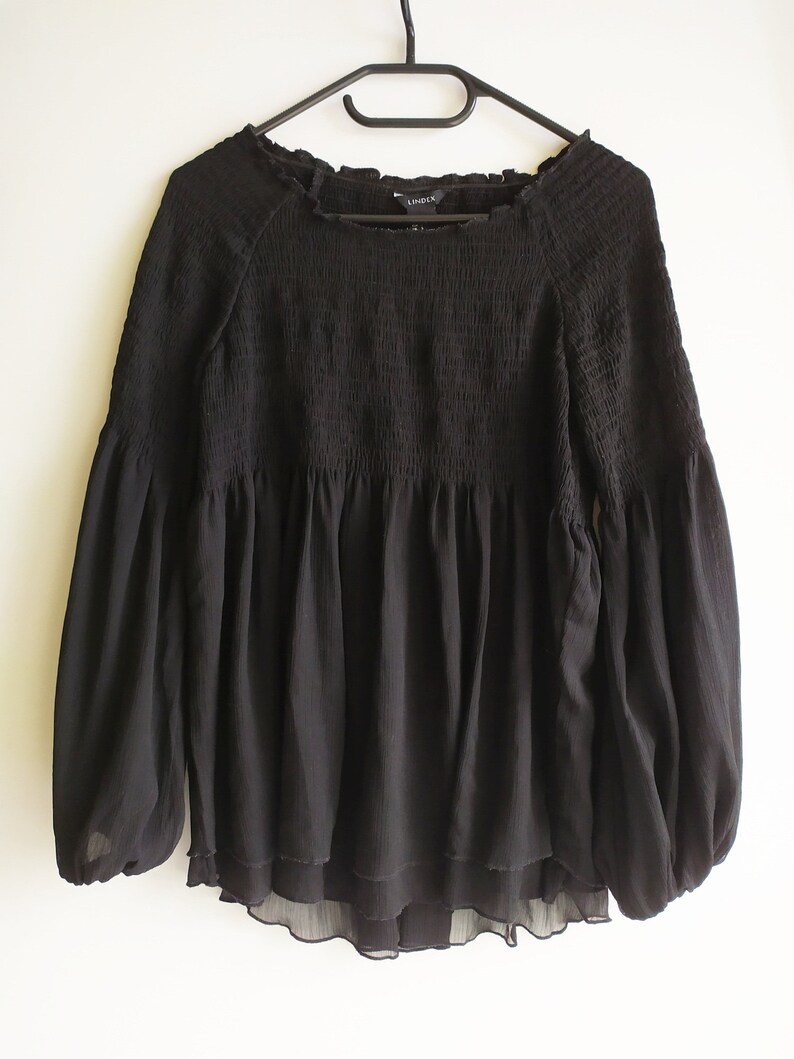 May include: Black long-sleeve blouse with a gathered, ruffled neckline and a tiered, ruffled hem. The blouse has a relaxed fit and is made of a lightweight, sheer fabric.