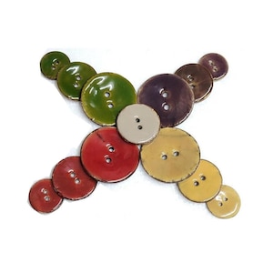 Könnte beinhalten: Eine Sammlung bunter Keramikknöpfe in verschiedenen Größen und Formen, die in einem Kreuzmuster angeordnet sind. Die Knöpfe sind in den Farben Grün, Rot, Gelb, Lila und Beige.