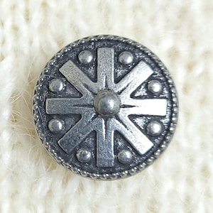 Op de afbeelding: Een ronde, zilverkleurige metalen knoop met een verhoogd achtpuntig stervormig ontwerp in het midden. De knoop heeft een textuurrijke, kralenrand en kleine verhoogde stippen tussen de punten van de ster, wat een gedetailleerd reliëfpatroon vormt.