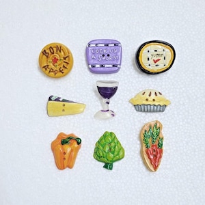 Könnte beinhalten: Sortiment von zehn bunten Keramikknöpfen mit verschiedenen Essens- und Getränkemotiven. Die Knöpfe zeigen Bilder von einem Weinglas, einem Stück Käse, einer Torte, einer Paprika, einer Artischocke, Karotten, einem Teller mit dem Wort "yumm", einem Teller mit den Worten "Bon Appetit" und einem Teller mit den Worten "Cook up a storm".