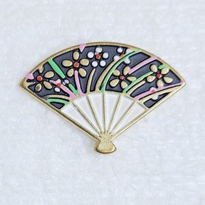 May include: A gold-colored metal fan with a black and white design. The fan is decorated with pink, green, and white flowers.