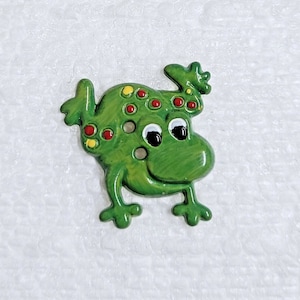 Könnte beinhalten: Ein grüner Froschknauf mit roten und gelben Flecken und weißen Augen. Der Frosch lächelt und hat die Arme erhoben.