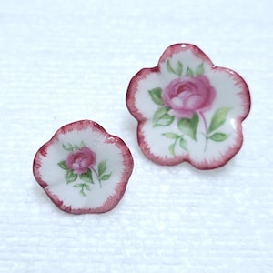 Puede incluir: Dos pendientes de porcelana con forma de flor. Cada pendiente presenta una base blanca con una rosa rosa y hojas verdes. Los bordes están ribeteados con un borde rosa oscuro. Un pendiente es más pequeño que el otro.