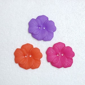 Puede incluir: Tres botones de plástico con forma de flor en morado, naranja y rosa. Cada botón tiene dos agujeros en el centro.