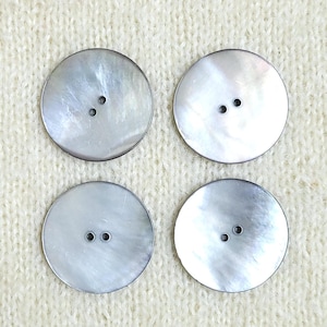 Puede incluir: Cuatro botones circulares con un acabado liso e iridiscente en tonos gris claro y plateado. Cada botón presenta dos pequeños orificios centrados para su fijación, mostrando un brillo nacarado natural en las superficies planas y redondas.