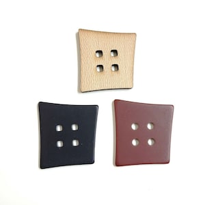 Może przedstawiać: Trzy kwadratowe guziki z czterema otworami każdy. Guziki są wykonane z drewna, czarnego plastiku i czerwonego plastiku.