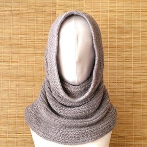 May include: A gray knitted cowl neck scarf with a hood. The scarf is folded and draped over a mannequin.