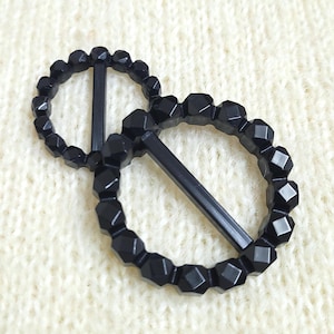 May include: Two circular black buckles with a faceted, bead-like texture along the outer rim. Each buckle features a single straight horizontal bar across the centre, designed for securing fabric or straps through the open circular frame.