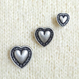 May include: Three heart-shaped buttons in varying sizes, each featuring a raised, smooth silver-toned centre surrounded by a dark, recessed border with intricate, embossed scrollwork patterns along the outer edge of the heart shape.