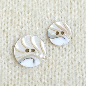 May include: Two round, flat buttons featuring a pearlescent, iridescent finish with swirling cream, beige, and white patterns. Each button has two centered holes for attachment and a smooth, polished surface with subtle dark veining details.