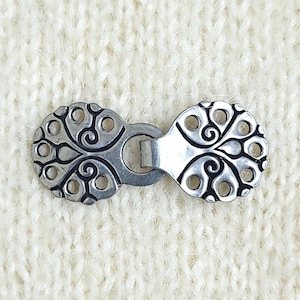 May include: A silver-toned metal cloak clasp featuring two circular components with intricate cut-out patterns and black recessed detailing. The two pieces are connected by a central metal loop, creating a functional fastener for garments.