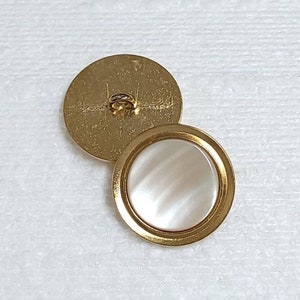 以下が含まれることがあります： 白蝶貝の中央に丸い金色のボタンが2つあります。左側のボタンは裏側にシャンクがあり、右側のボタンは裏側に平らな面があります。