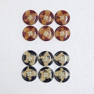 Könnte beinhalten: Eine Sammlung von zwölf runden Knöpfen. Sechs Knöpfe haben ein braunes und beiges Schildpattmuster, und sechs haben ein schwarzes und beiges Schildpattmuster. Jeder Knopf hat vier Löcher zum Annähen.