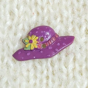A colourful novelty button which is shaped like a purple rimmed hat with a yellow flower. The button has two holes for sewing and is shown on a cream-coloured knitted background.