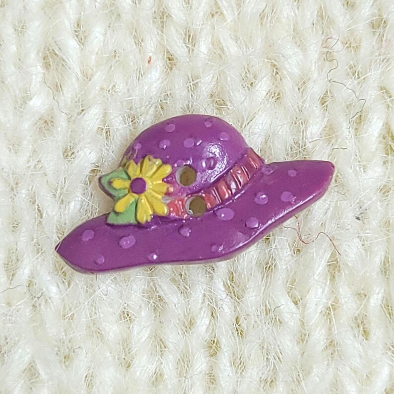 A colourful novelty button which is shaped like a purple rimmed hat with a yellow flower. The button has two holes for sewing and is shown on a cream-coloured knitted background.