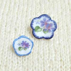 May include: Two flower-shaped ceramic buttons with a white base, featuring hand-painted purple blossoms, green leaves, and a blue-rimmed edge. The larger button has a dark blue, feathered border, while the smaller button has a solid blue outline.