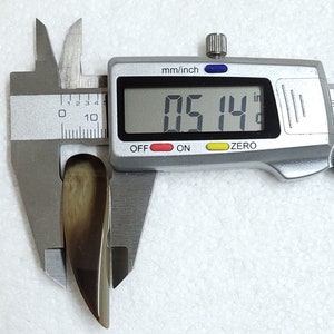 May include: A polished, teardrop-shaped object in shades of brown and tan is measured with a digital caliper. The measurement reads 0.514 inches. The caliper is silver, and the object appears to be made of a natural material.