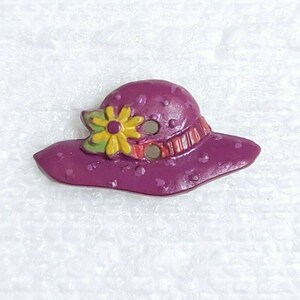 May include: A purple and white polka dot sun hat with a yellow flower and red and orange stripes. The hat is a decorative button.