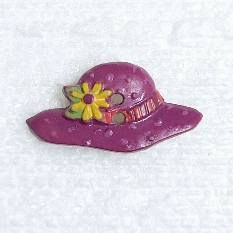 May include: A purple and white polka dot sun hat with a yellow flower and red and orange stripes. The hat is a decorative button.