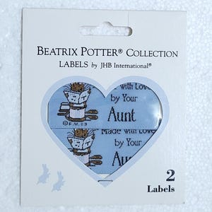 May include: Beatrix Potter Collection labels by JHB International. The package features a heart-shaped window revealing two blue labels with a repeating illustration and the text "with Love by Your Aunt". The package has a white background and includes the number "2 Labels".
