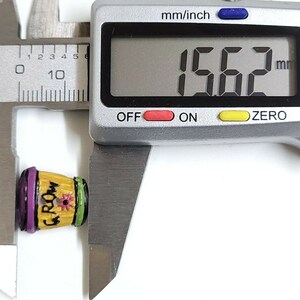 May include: A small, yellow, wooden, decorative pot with a purple rim and green trim. The pot has the word "Groom" written on it and a small flower painted on the front. The pot is being measured with a digital caliper, which reads 15.62 mm.