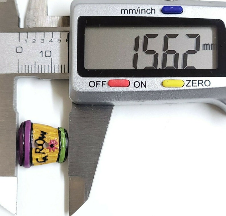May include: A small, yellow, wooden, decorative pot with a purple rim and green trim. The pot has the word "Groom" written on it and a small flower painted on the front. The pot is being measured with a digital caliper, which reads 15.62 mm.