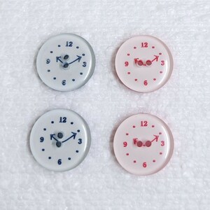 May include: Four round, plastic buttons with a clock face design. Two buttons are white with blue hands and numbers, and two buttons are pink with red hands and numbers.