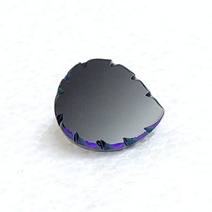 Puede incluir: Una piedra preciosa negra con una forma facetada irregular. La piedra preciosa tiene un brillo iridiscente púrpura y verde.