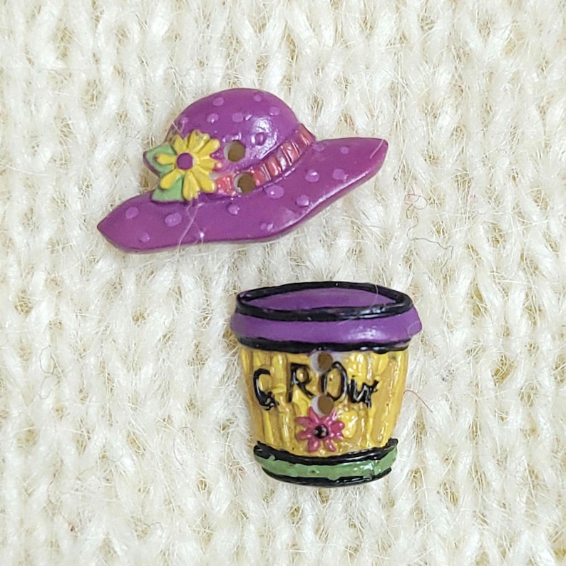 Two colourful buttons, one shaped like a purple rimmed hat with a yellow flower and the other shaped like a yellow flower pot with the word "GROW" on it. Each button has two holes for sewing.  They are arranged on a cream-coloured knitted background.