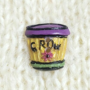 A colourful novelty button shaped like a yellow flower pot with the word "GROW" on it. The button has two holes for sewing and is shown on a cream-coloured knitted background.