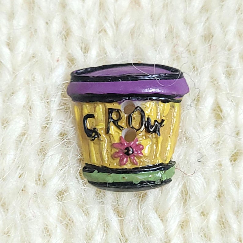 A colourful novelty button shaped like a yellow flower pot with the word "GROW" on it. The button has two holes for sewing and is shown on a cream-coloured knitted background.