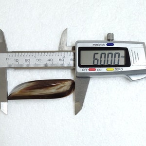 May include: A polished, elongated gemstone with a mix of brown and cream colors. The digital caliper shows the stone measures 60.00 mm in length, approximately 2.36 inches. The stone has a smooth, curved shape.