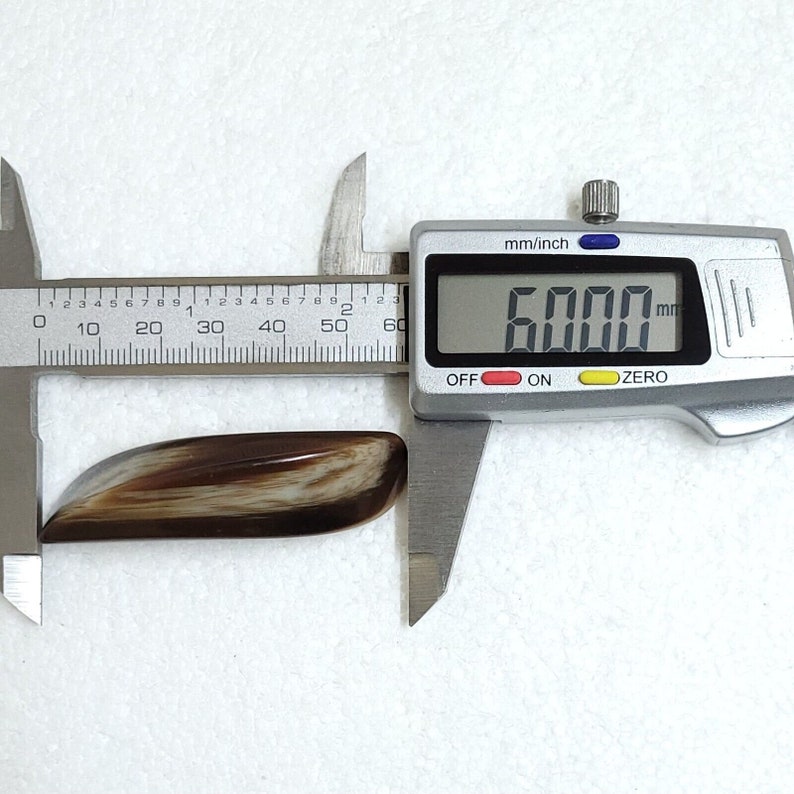 May include: A polished, elongated gemstone with a mix of brown and cream colors. The digital caliper shows the stone measures 60.00 mm in length, approximately 2.36 inches. The stone has a smooth, curved shape.