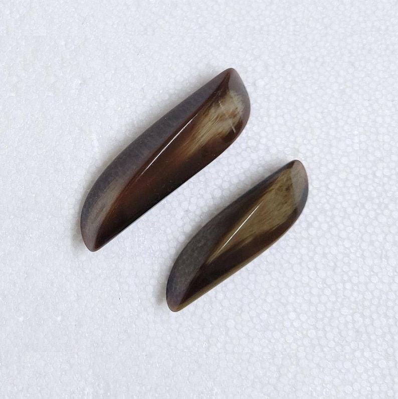 May include: Two polished, elongated gemstones with a smooth, glossy finish. The stones are a blend of brown and tan hues, with a subtle gradient effect. The shape is reminiscent of a leaf or a teardrop.