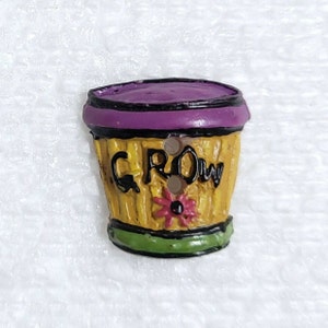 May include: A yellow ceramic button shaped like a flower pot with a purple rim and the word "GROW" written on the side. The pot has a green base and a pink flower on the front.