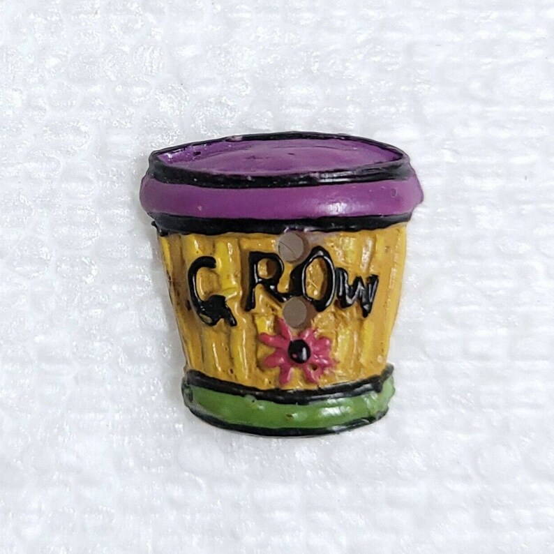 May include: A yellow ceramic button shaped like a flower pot with a purple rim and the word "GROW" written on the side. The pot has a green base and a pink flower on the front.
