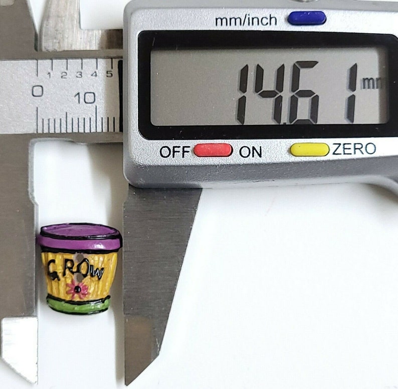 May include: A small, yellow ceramic planter with a purple rim and the word "GROW" written on it. The planter is 1.46 inches in diameter.