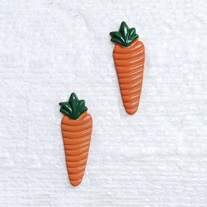 Puede incluir: Dos adornos en forma de zanahoria naranja con copas verdes y frondosas. Las zanahorias tienen una superficie texturizada con líneas verticales. El fondo es una superficie blanca y texturizada. Estos artículos decorativos podrían usarse para manualidades o joyería.