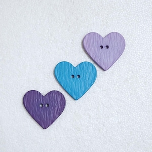 May include: Three heart-shaped buttons in shades of purple and blue. Each button has two holes for sewing. The buttons have a textured, wood-like appearance, and are arranged diagonally on a white surface.