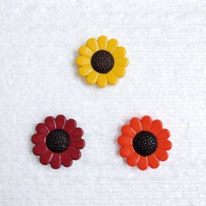 May include: Three plastic sunflower buttons. One is yellow, one is red, and one is orange. Each button has a black centre.