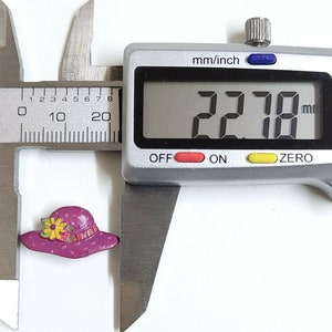 May include: A close-up of a purple plastic button shaped like a hat with a yellow flower and white polka dots. The button is being measured with a caliper, which reads 22.78 mm.