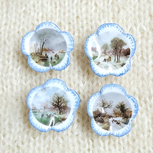 May include: Four flower-shaped ceramic buttons with a blue-painted scalloped edge. Each button features a unique, detailed landscape scene in the center, depicting trees, open fields, and small figures in a muted, natural color palette.