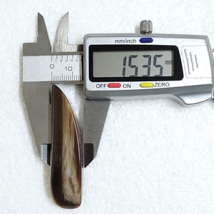 May include: A polished, elongated object with a brown and cream-colored striped pattern, measuring 15.35 mm in length, is held by a digital caliper. The caliper displays the measurement in millimeters and inches.