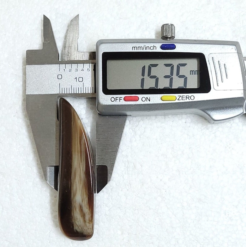 May include: A polished, elongated object with a brown and cream-colored striped pattern, measuring 15.35 mm in length, is held by a digital caliper. The caliper displays the measurement in millimeters and inches.