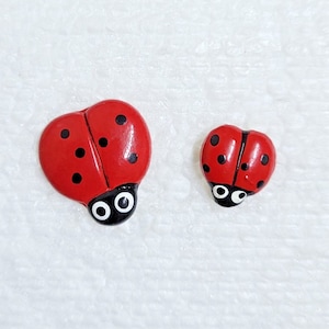 May include: Two red ladybug shaped buttons with black spots and white eyes. The larger ladybug is about 2.5 cm in diameter and the smaller ladybug is about 1.25 cm in diameter.